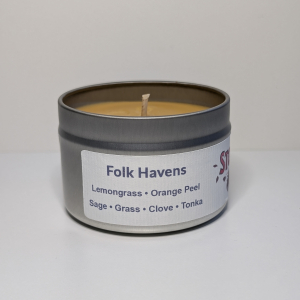 A photo of a small round silver tin containing a 4 oz soy wax candle with a short wick. The candle wax is warm yellow in color, and a white label on the tin includes a list of the scent notes, a QR code, logo, and social handle for The Storyteller Squad, and the name of the candle, all in front of the silhouettes of a group of evergreen trees.