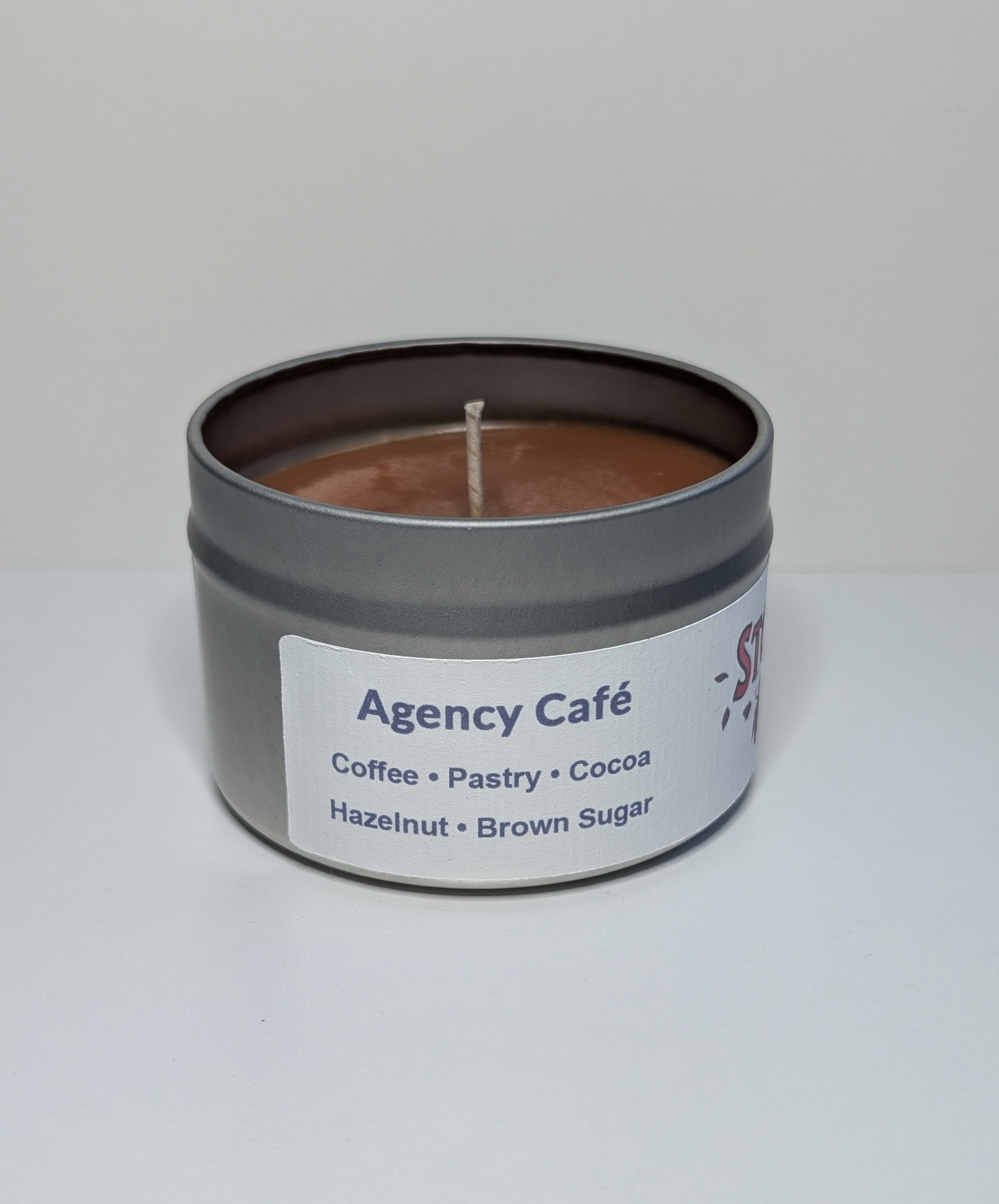 A photo of a small round silver tin containing a 4 oz soy wax candle with a short wick. The candle wax is brown in color, and a white label on the tin includes a list of the scent notes, a QR code, logo, and social handle for The Storyteller Squad, and the name of the candle, all in front of the silhouettes of a group of evergreen trees.