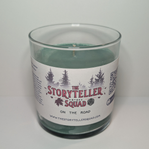 A photo of a glass tumbler with a bamboo lid containing a 10 oz soy wax candle with a short wick. The candle wax is dark green in color, and a white label on the glass lists the scent notes, a short section of copy about the candle's scent profile, a QR code, website address, and logo for The Storyteller Squad, and the name of the candle, all in front of the silhouettes of a group of evergreen trees.