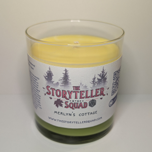 A photo of a glass tumbler with a bamboo lid containing a 10 oz soy wax candle with a short wick. The candle wax is bright green in color, and a white label on the glass lists the scent notes, a short section of copy about the candle's scent profile, a QR code, website address, and logo for The Storyteller Squad, and the name of the candle, all in front of the silhouettes of a group of evergreen trees.