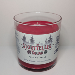 A photo of a glass tumbler with a bamboo lid containing a 10 oz soy wax candle with a short wick. The candle wax is bright red in color, and a white label on the glass lists the scent notes, a short section of copy about the candle's scent profile, a QR code, website address, and logo for The Storyteller Squad, and the name of the candle, all in front of the silhouettes of a group of evergreen trees.