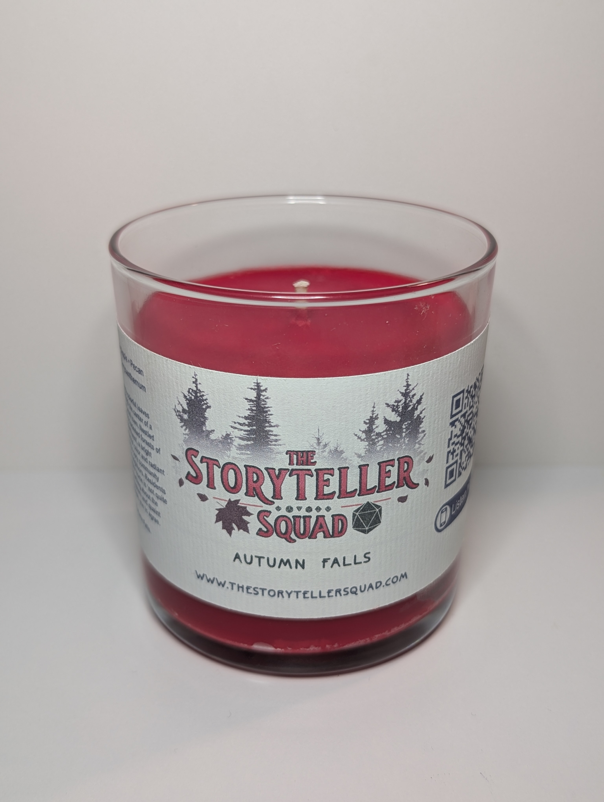 A photo of a glass tumbler with a bamboo lid containing a 10 oz soy wax candle with a short wick. The candle wax is bright red in color, and a white label on the glass lists the scent notes, a short section of copy about the candle's scent profile, a QR code, website address, and logo for The Storyteller Squad, and the name of the candle, all in front of the silhouettes of a group of evergreen trees.