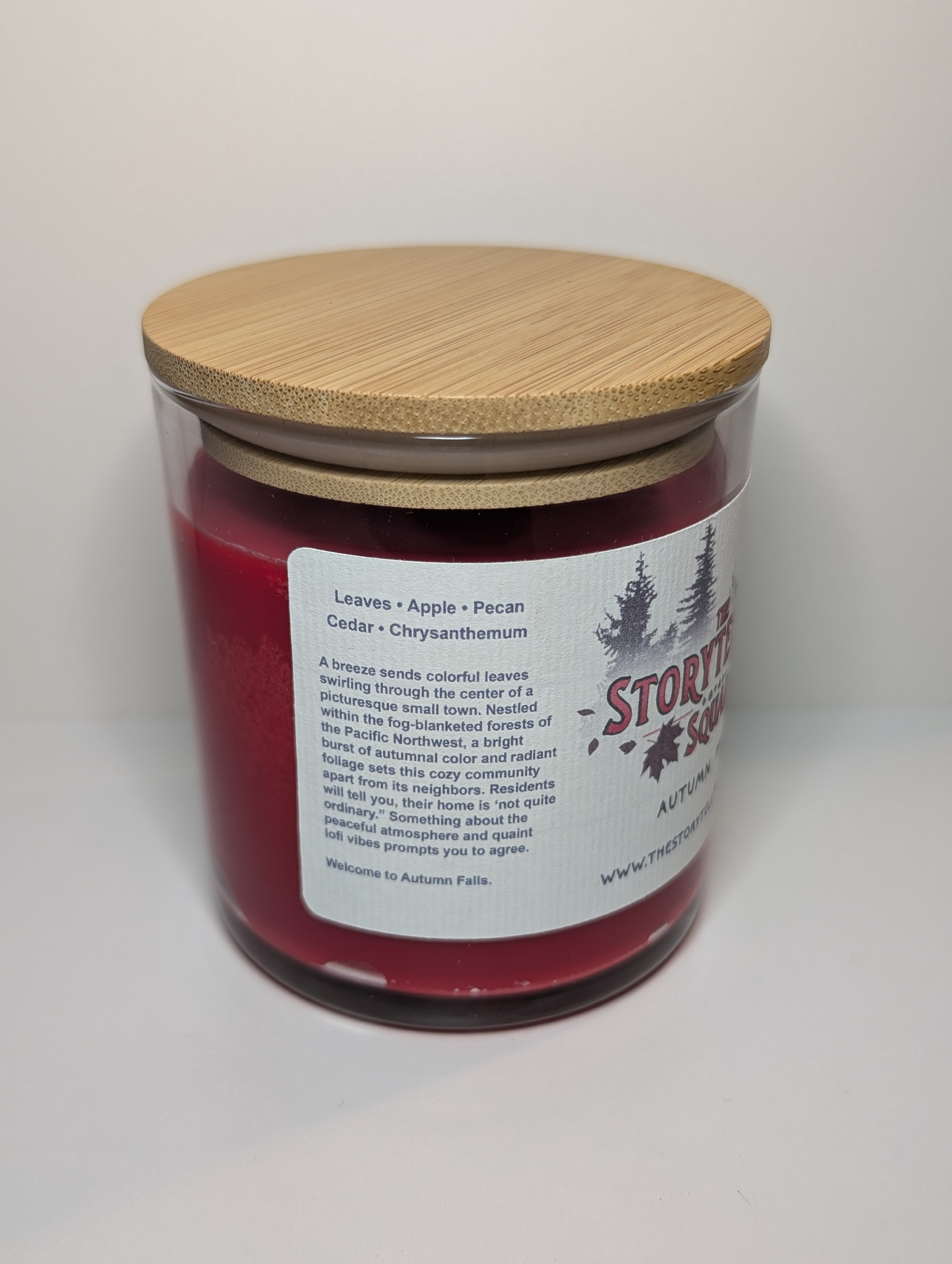 A photo of a glass tumbler with a bamboo lid containing a 10 oz soy wax candle with a short wick. The candle wax is bright red in color, and a white label on the glass lists the scent notes, a short section of copy about the candle's scent profile, a QR code, website address, and logo for The Storyteller Squad, and the name of the candle, all in front of the silhouettes of a group of evergreen trees.